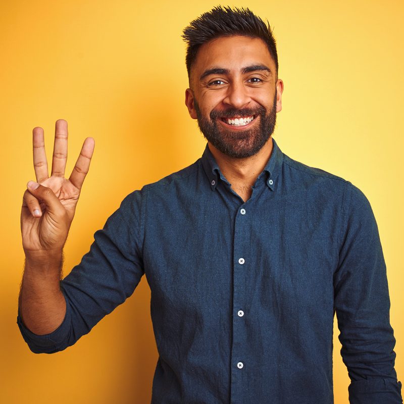 Young indian businessman wearing elegant shirt standing over isolated white background showing and pointing up with fingers number three while smiling confident and happy.